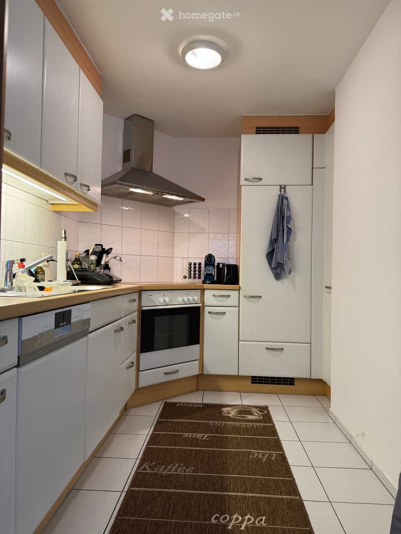 Modern kitchen with white cabinets, stainless steel appliances, and a tiled floor. Features include a refrigerator, oven, dishwasher, sink, and a coffee machine on the counter.