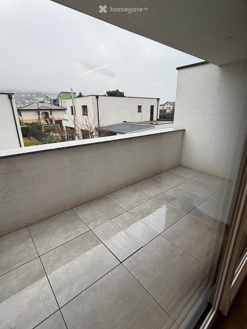 Empty balcony with tiled floor and low concrete railing, overlooking a residential area with houses and trees.
