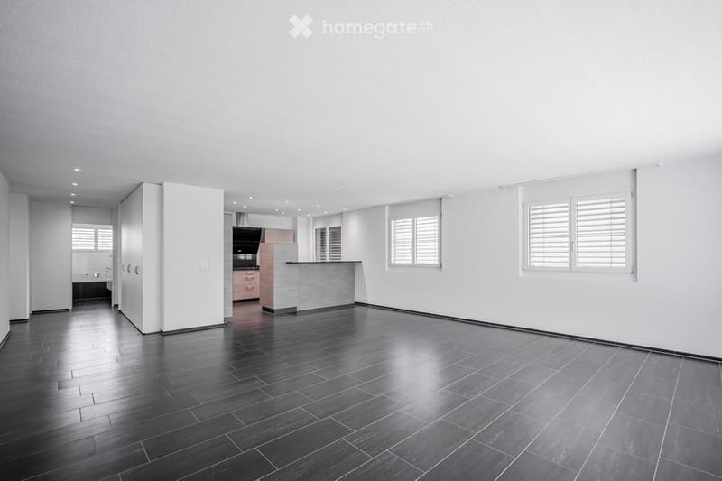 Empty room with dark wooden flooring, white walls, and large windows with shutters. The room features a modern design with recessed lighting and a visible kitchen area in the background.