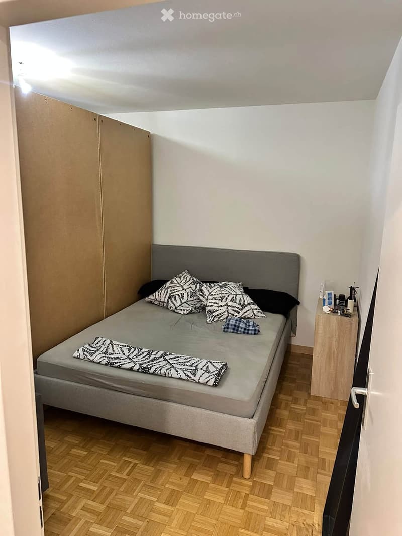 A small bedroom with a single bed, nightstand, and wardrobe. The bed has patterned pillows and a blanket, and the room features wooden flooring.