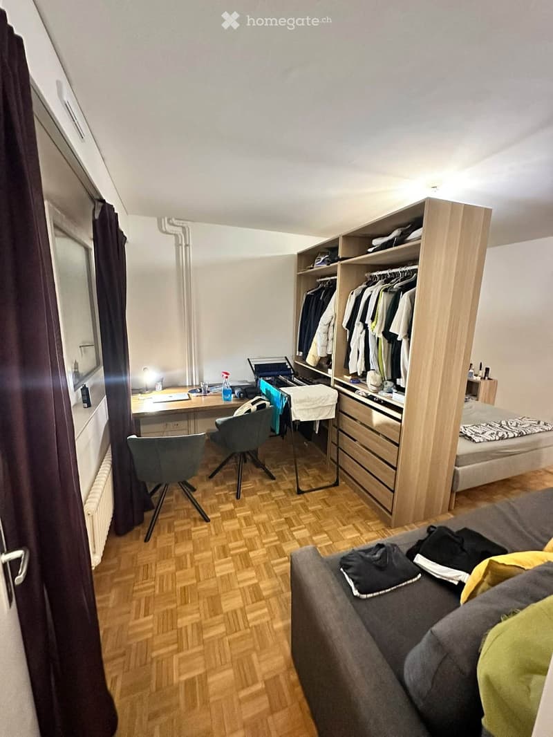 A compact, furnished room with a bed, sofa, desk, chair, wardrobe, and shelves. The room features wooden flooring and large windows with dark curtains.