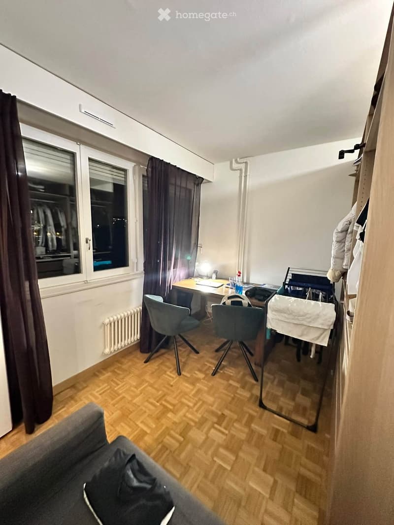 A small, furnished room with a sofa, desk, chairs, and a window with curtains. The room features wooden flooring and a radiator under the window.