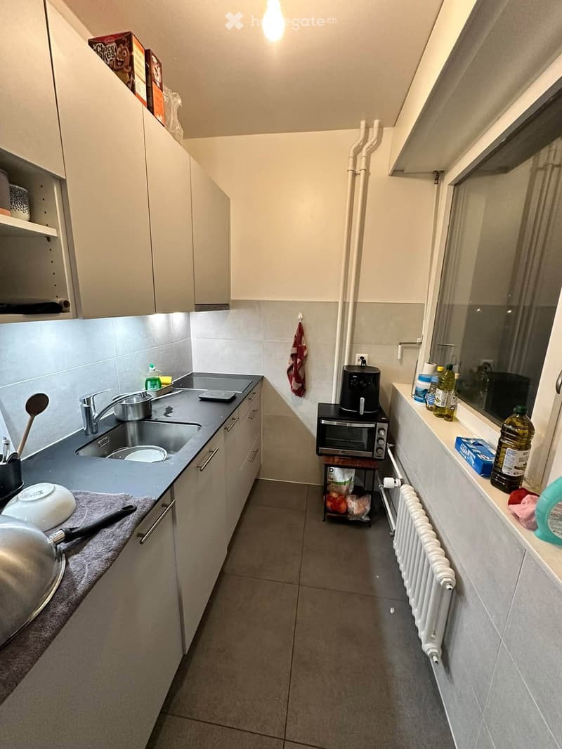 Modern kitchen with white cabinets, stainless steel sink, oven, and various appliances. Countertops hold a few items including a bottle, a cup, and a small basket with fruits.