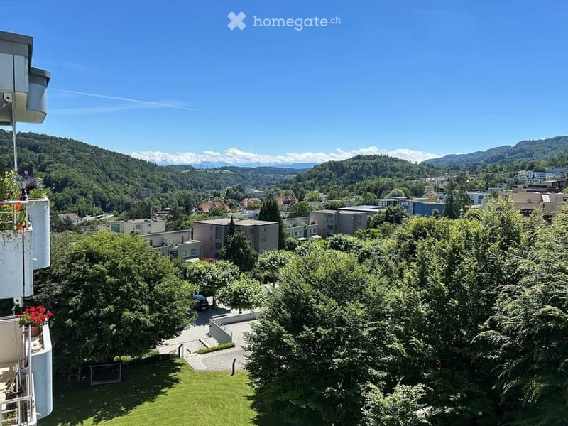 Balcony with a panoramic view of a cityscape, surrounded by greenery and trees, with distant mountains visible under a clear blue sky.