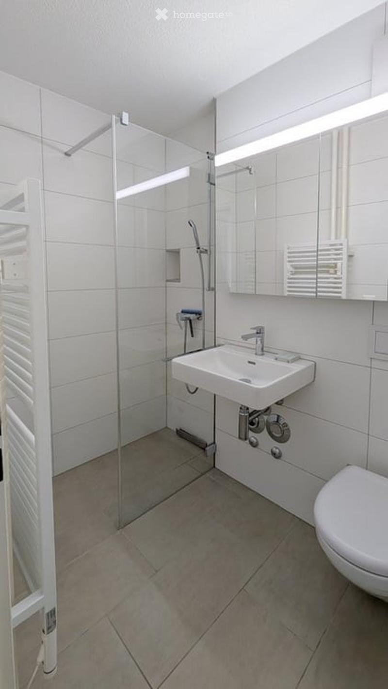 Modern bathroom with white tiles, a glass-enclosed shower, a sink with a mirror, and a toilet.