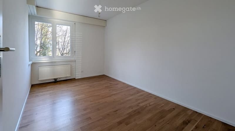 Empty room with wooden flooring, white walls, large window with blinds, radiator, and a door handle visible.