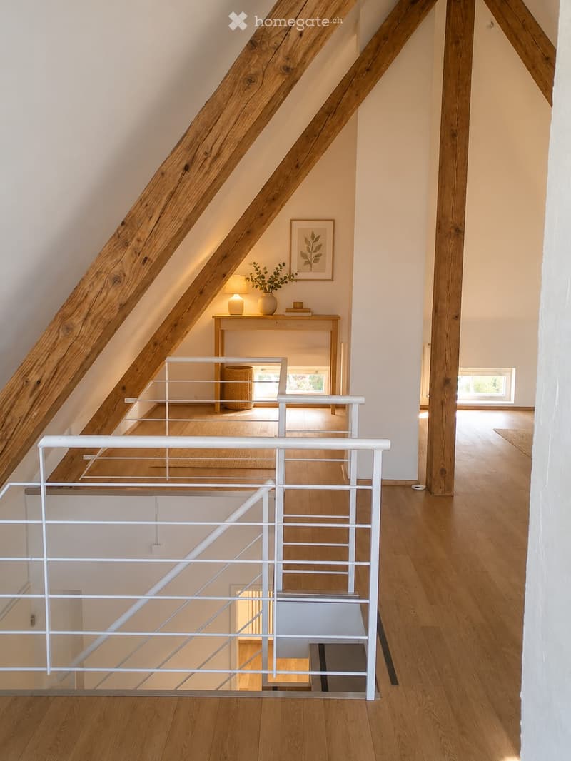 Modern hallway with wooden floors, white walls, and a staircase featuring a glass railing and wooden beams. A small table with a lamp and vase is visible in the background.
