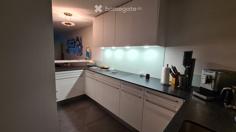 Modern kitchen with white cabinets, black countertops, and various appliances including a coffee maker and toaster. The space features a sleek design with integrated lighting and a clean, organized layout.