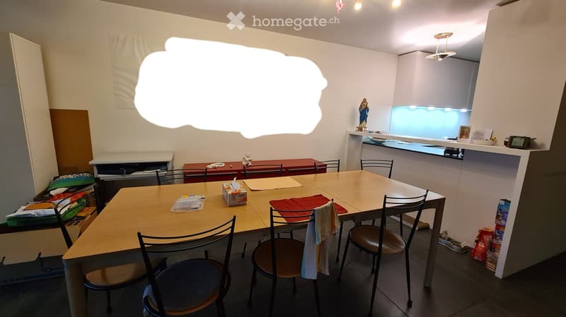 A furnished room with a dining table and chairs, a kitchen area with a counter, and various items on the counter and shelves.