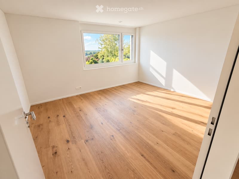 Bright, empty room with wooden flooring and large windows offering a view of greenery outside.