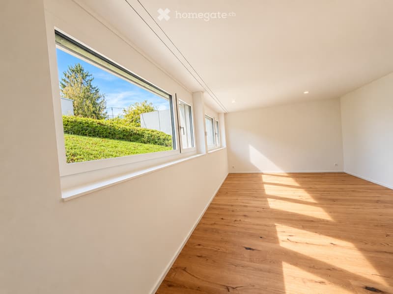 Empty room with light-colored walls and wooden flooring, featuring large windows that let in natural light and offer a view of a green outdoor area.
