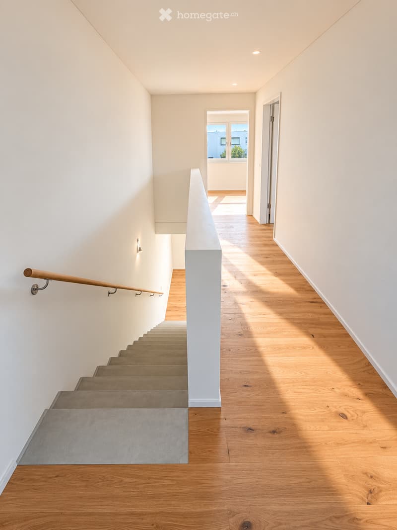 Modern hallway with wooden flooring, white walls, and a staircase leading upwards. Features a wooden handrail and recessed lighting.