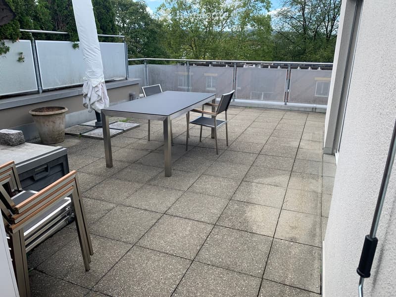 Balcony with tiled floor, metal table, and chairs, featuring a view of greenery and a covered umbrella.