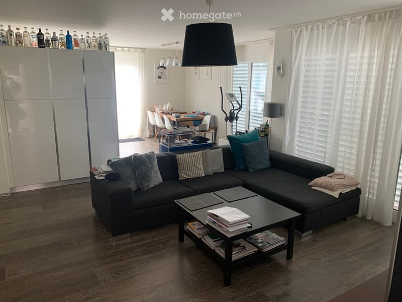 A modern living room with a grey sectional sofa, coffee table, exercise bike, dining area with chairs and table, and a refrigerator with bottles on top.