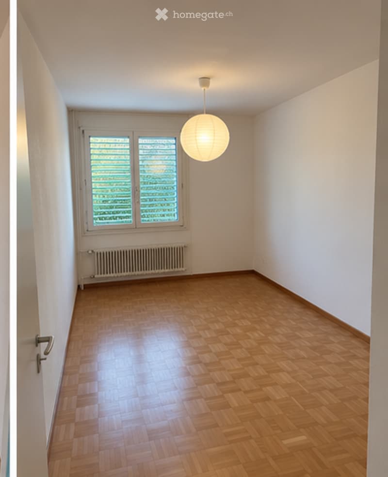 Empty room with wooden parquet flooring, white walls, a single window with blinds, a radiator, and a hanging spherical light fixture.
