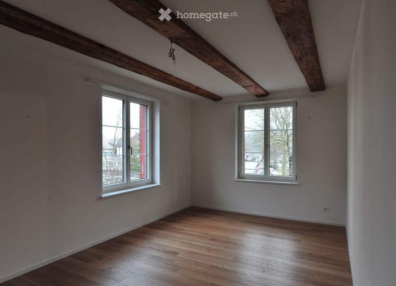 Empty room with wooden flooring, exposed wooden beams on the ceiling, and two windows offering natural light.