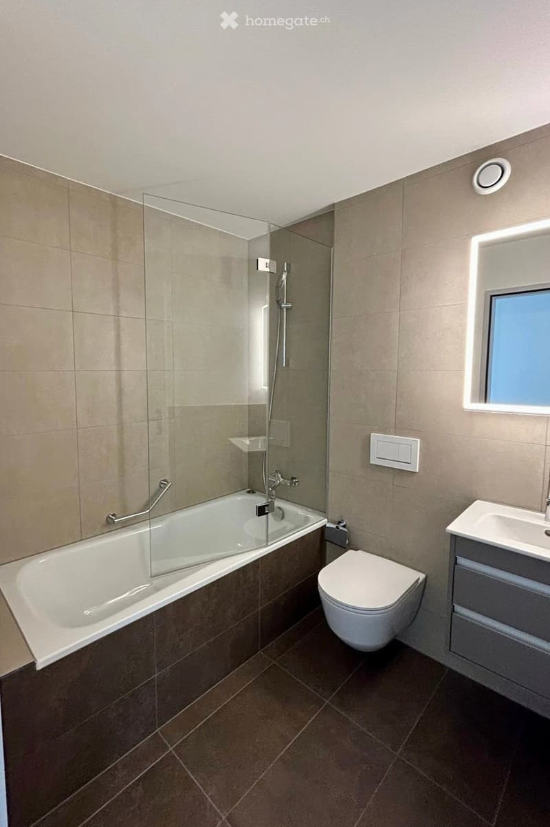 Modern bathroom with a bathtub, toilet, sink, and a large mirror with built-in lighting. The space features beige tiles and a glass-enclosed shower area.