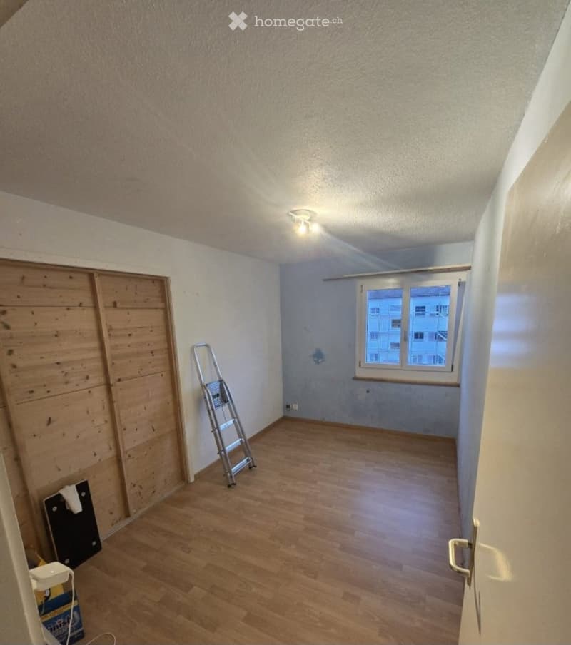 Empty room with wooden flooring, white walls, and a window with a view of buildings outside. A ladder and some construction materials are present.