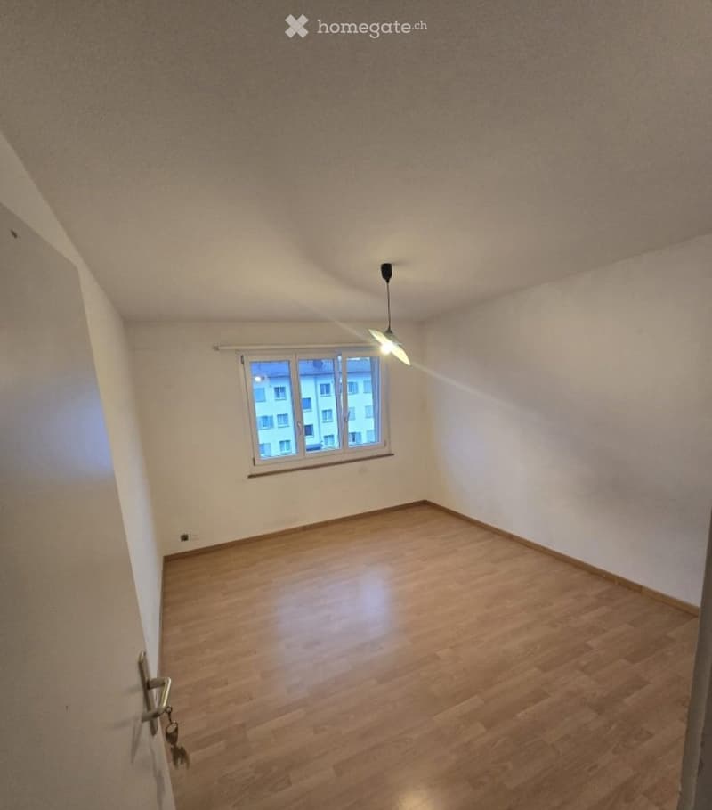 Empty room with wooden flooring, white walls, and a single window with a view of a building outside. The room has a simple ceiling light fixture and a door with a handle visible on the left side.