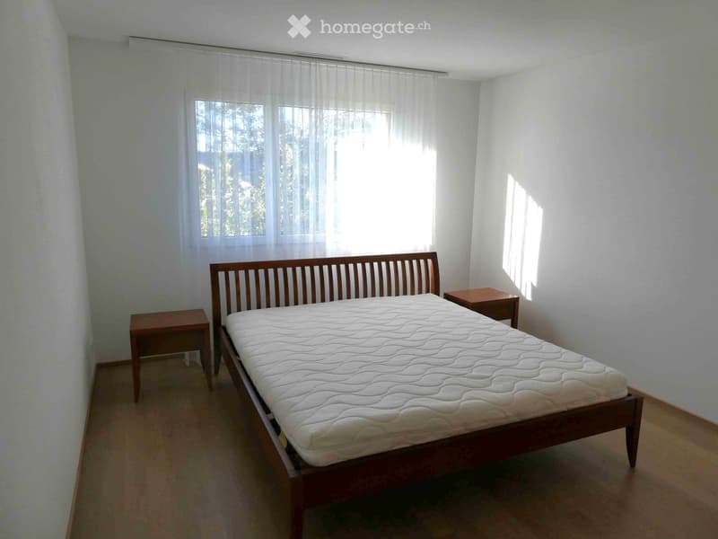 A well-lit bedroom with a wooden bed, white mattress, and two wooden nightstands, featuring sheer curtains on the windows.