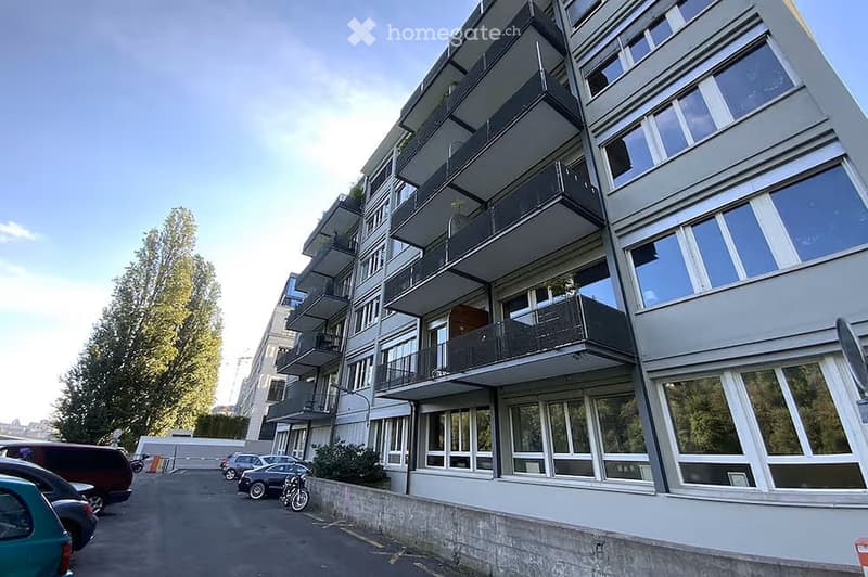 Modern multi-story apartment building with large windows and balconies, surrounded by parked cars and greenery.
