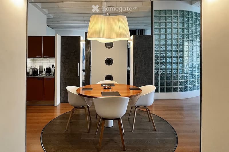 Modern dining area with a round wooden table and white chairs, adjacent to a kitchen with dark cabinets and stainless steel appliances.