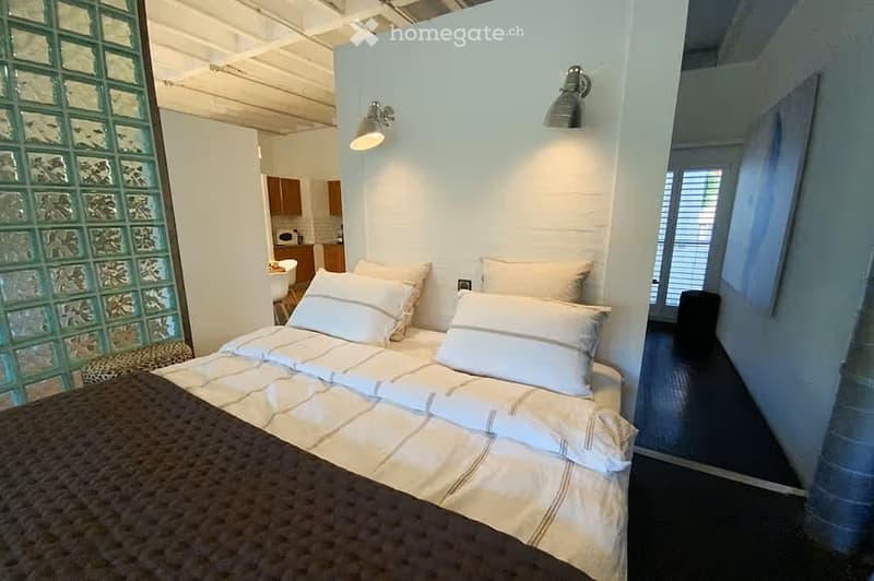 A modern bedroom with a neatly made bed, white pillows, and a nightstand. A small kitchen area is visible in the background with cabinets and a microwave. A glass block wall and a window with blinds add natural light.