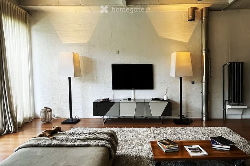 Modern living room with wooden flooring, white brick walls, large windows with curtains, a sofa with a fur throw, a coffee table with books, two floor lamps, a TV on a sleek stand, and a decorative basket on the floor.
