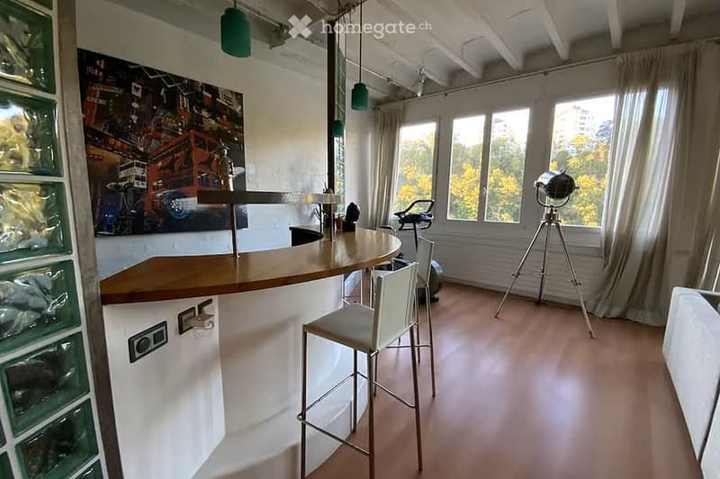 Modern, open-plan room with a bar counter, two stools, large windows with curtains, and a tripod with a camera in the corner.