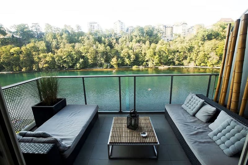 Balcony with a view of a river and trees, featuring a sofa, a small table, a lantern, and a potted plant.