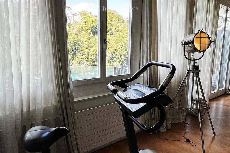 A room with a treadmill and a vintage-style lamp on a tripod stand, large windows with sheer curtains, and a view of greenery outside.