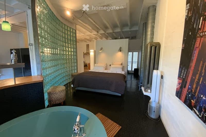 Modern bedroom with a large bed, a small stool, a bathtub, and a large picture frame on the wall. The room features exposed ceiling beams and a glass partition separating the sleeping area from the bathroom.