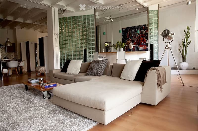 Modern living room with a large sectional sofa, coffee table with books, pendant lights, and a view into a kitchen area with bar stools.