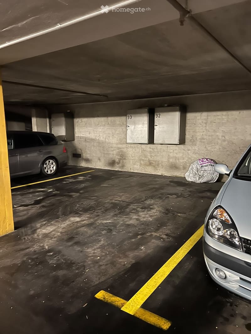 Underground parking area with two parked cars, marked parking spaces, and numbered storage units on the wall.