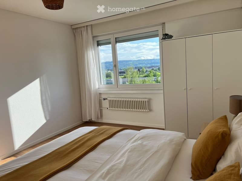 Bright bedroom with a large window offering a scenic view, featuring a neatly made bed with pillows and a blanket, a wardrobe, and a radiator below the window.