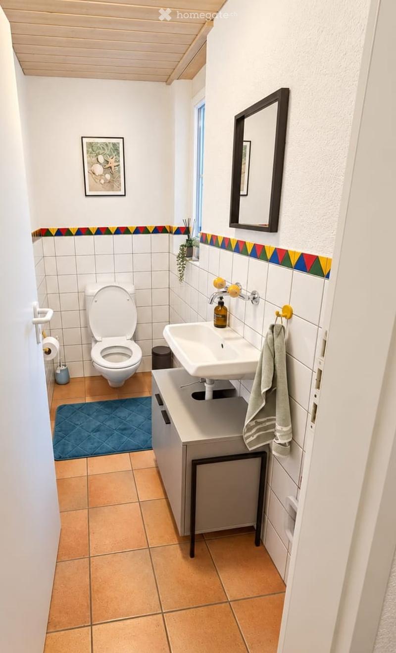 Cozy bathroom with white tiles, a toilet, a sink with a mirror above it, a small cabinet, a towel, and a decorative plant on the windowsill.