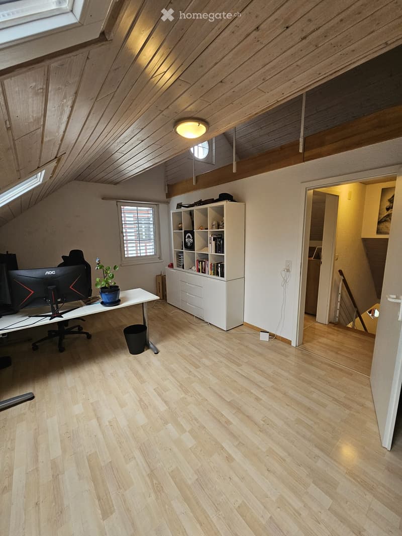 Bright attic office with wooden floors, slanted wooden ceiling, large windows, desk with computer, bookshelf, and small plant.