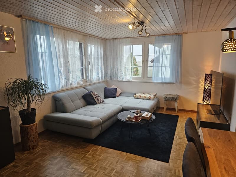 Modern living room with a grey sectional sofa, coffee table, wooden flooring, large windows with sheer curtains, and decorative items including a potted plant and a mirror.