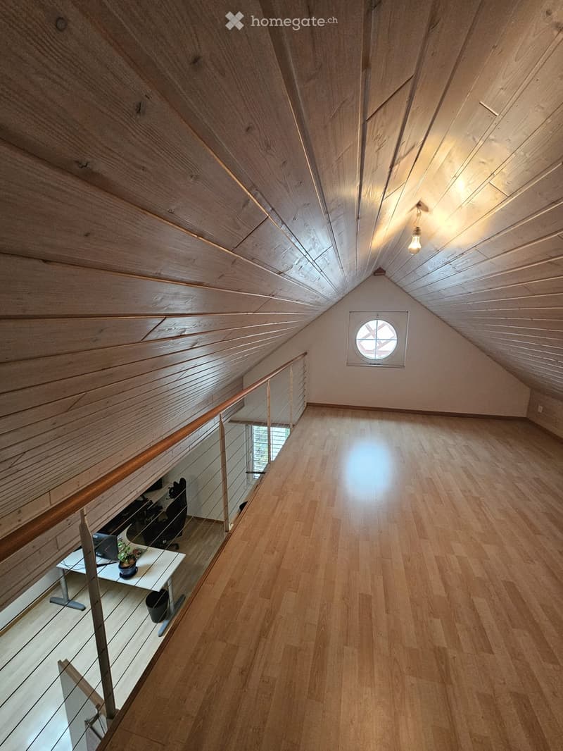 Spacious, well-lit room with wooden floors and slanted wooden ceilings. Features a modern glass railing leading to a lower level, a small desk with a computer and potted plant, and a round window near the ceiling.