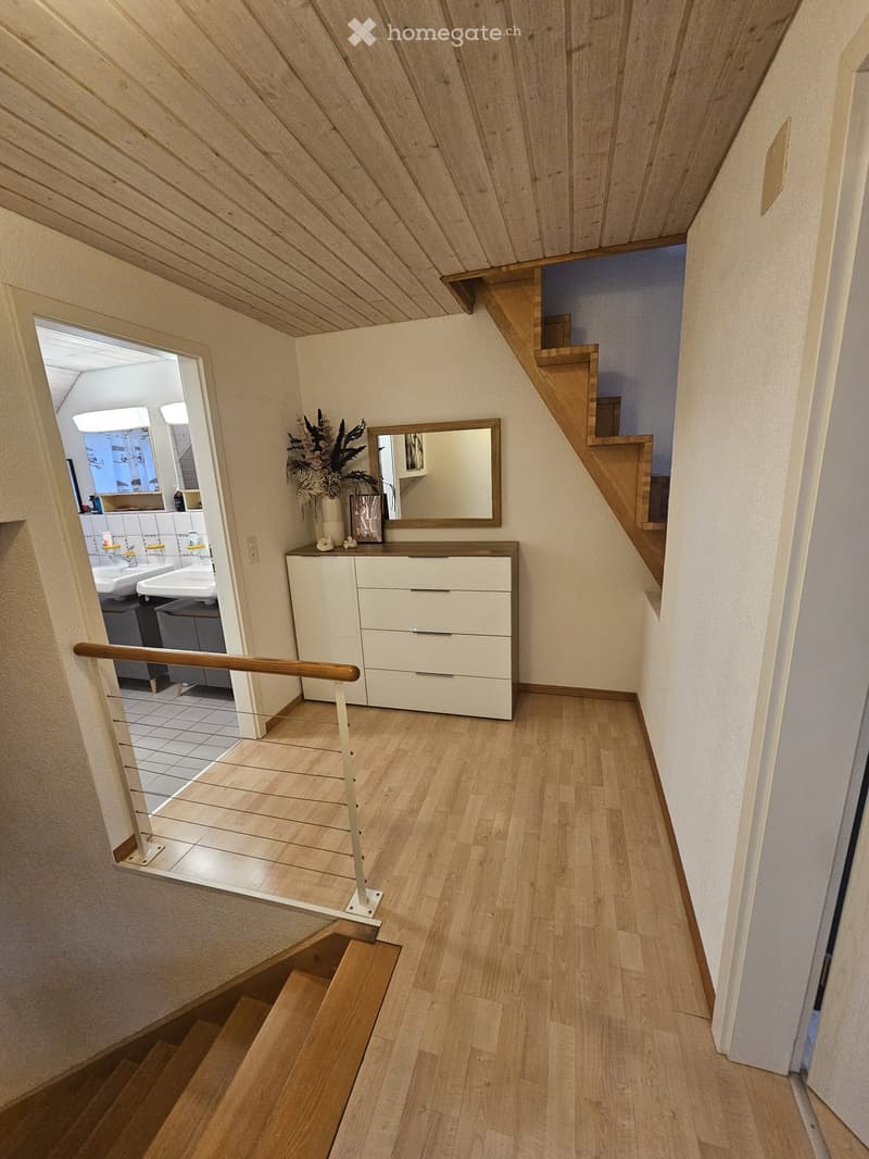 A narrow hallway with wooden flooring, a staircase leading upwards, a small white dresser with drawers, a mirror, and a vase with flowers. A bathroom is visible through an open doorway on the left.