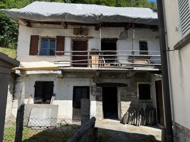 A rustic stone house with a covered balcony, wooden shutters, and a tarp-covered roof. The building shows signs of renovation with exposed wooden beams and a metal railing on the balcony.