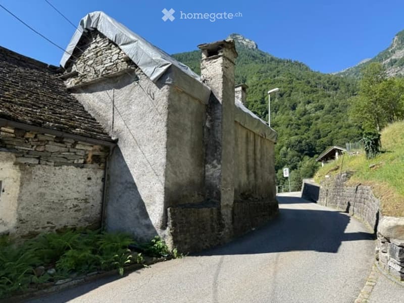 A rustic stone building with a tiled roof and chimney, situated on a narrow road surrounded by greenery and mountains.