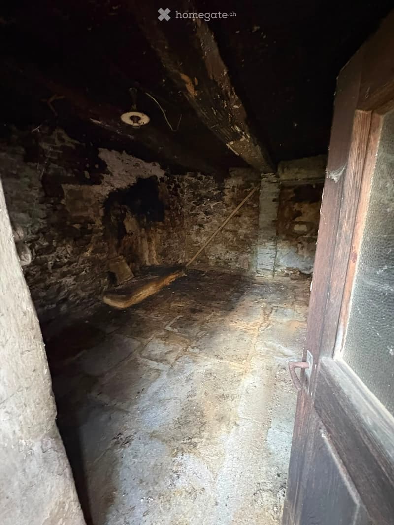 An unfurnished room with exposed stone walls, a wooden door frame, and a concrete floor. The space appears to be a basement or storage area with minimal furnishings.