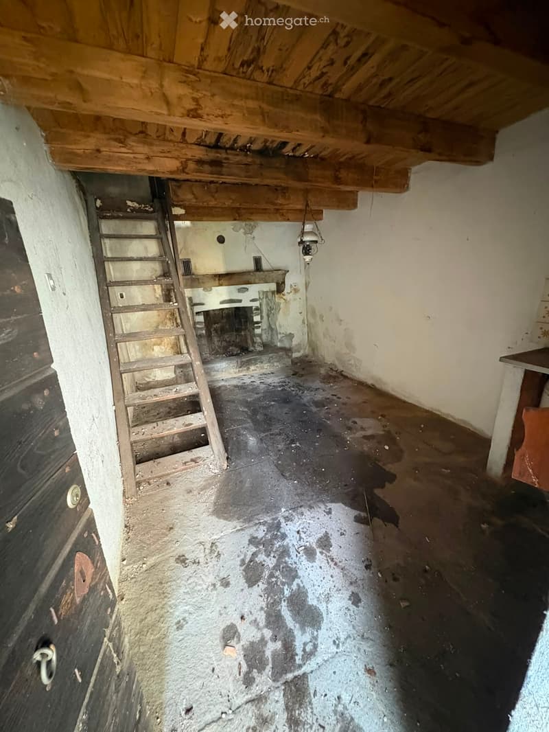 Empty room with exposed wooden beams on the ceiling, a wooden ladder leading to an attic, and a small table in the corner. The floor is concrete, and the walls are unfinished plaster.