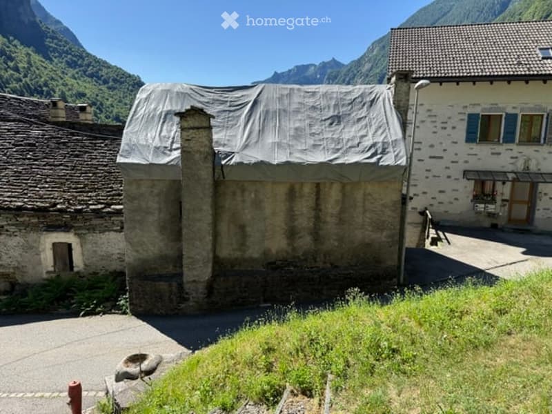 A rustic stone building with a tarp-covered roof, situated in a mountainous area with visible greenery and distant peaks.