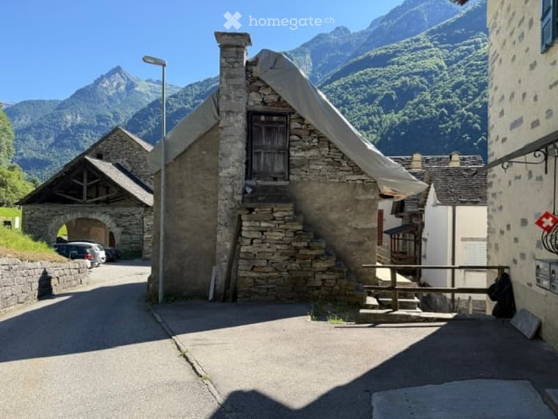 A rustic stone building with a slanted roof and wooden shutters, situated in a picturesque mountain village with clear blue skies and distant peaks.