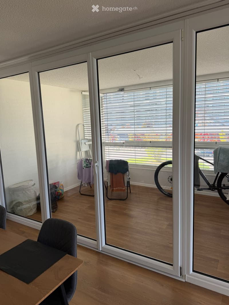 A modern, well-lit room with wooden flooring, large glass sliding doors leading to a balcony, and a view of the outside. The room features a black chair, a small wooden table, and a bicycle positioned near the balcony.