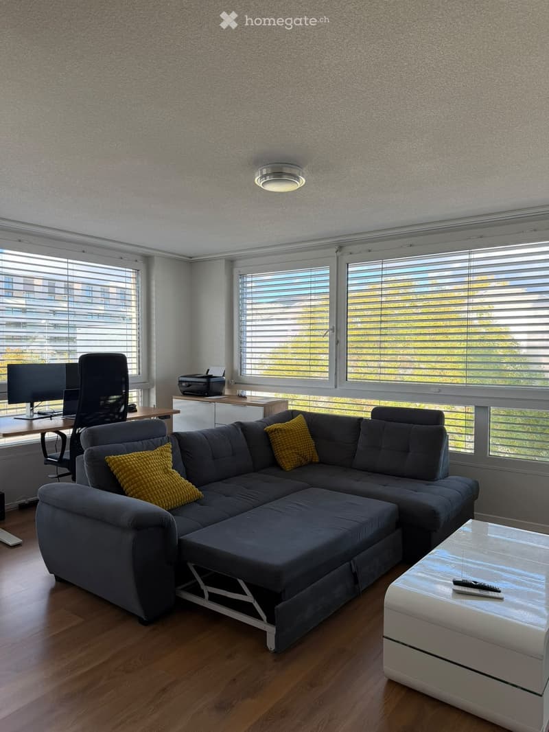 Modern living room with grey sectional sofa, yellow throw pillows, white coffee table, desk with chair, and large windows with blinds.
