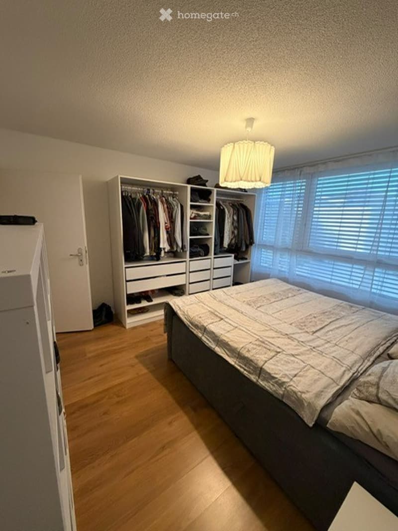 Well-lit bedroom with wooden flooring, a large bed with bedding, a wardrobe with clothes, and a ceiling lamp.