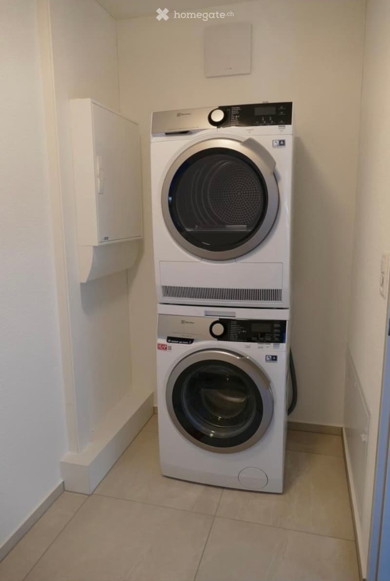 A compact bathroom with a stacked washing machine and dryer unit, beige tiled floor, and white walls.